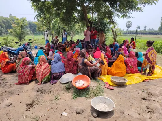 Workers sitting on the ground under trees for an outdoor meeting, with a field behind them.