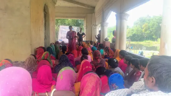 Women and men seated on the floor of a veranda at an awareness meeting.
