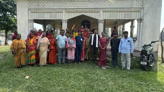 A group of women and men posed in front of a building beside a field.