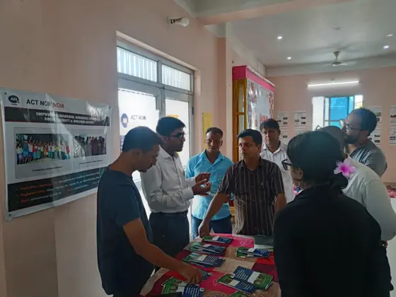 People looking at printed material at a table, with an Act Now India poster behind them.