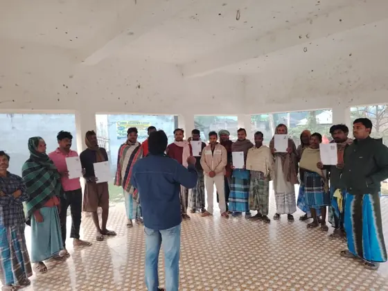 A man addressing workers who hold papers, in an open hall.