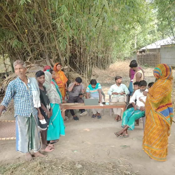 People seated at a table outdoors under bamboo, with a woman in a yellow sari standing nearby.